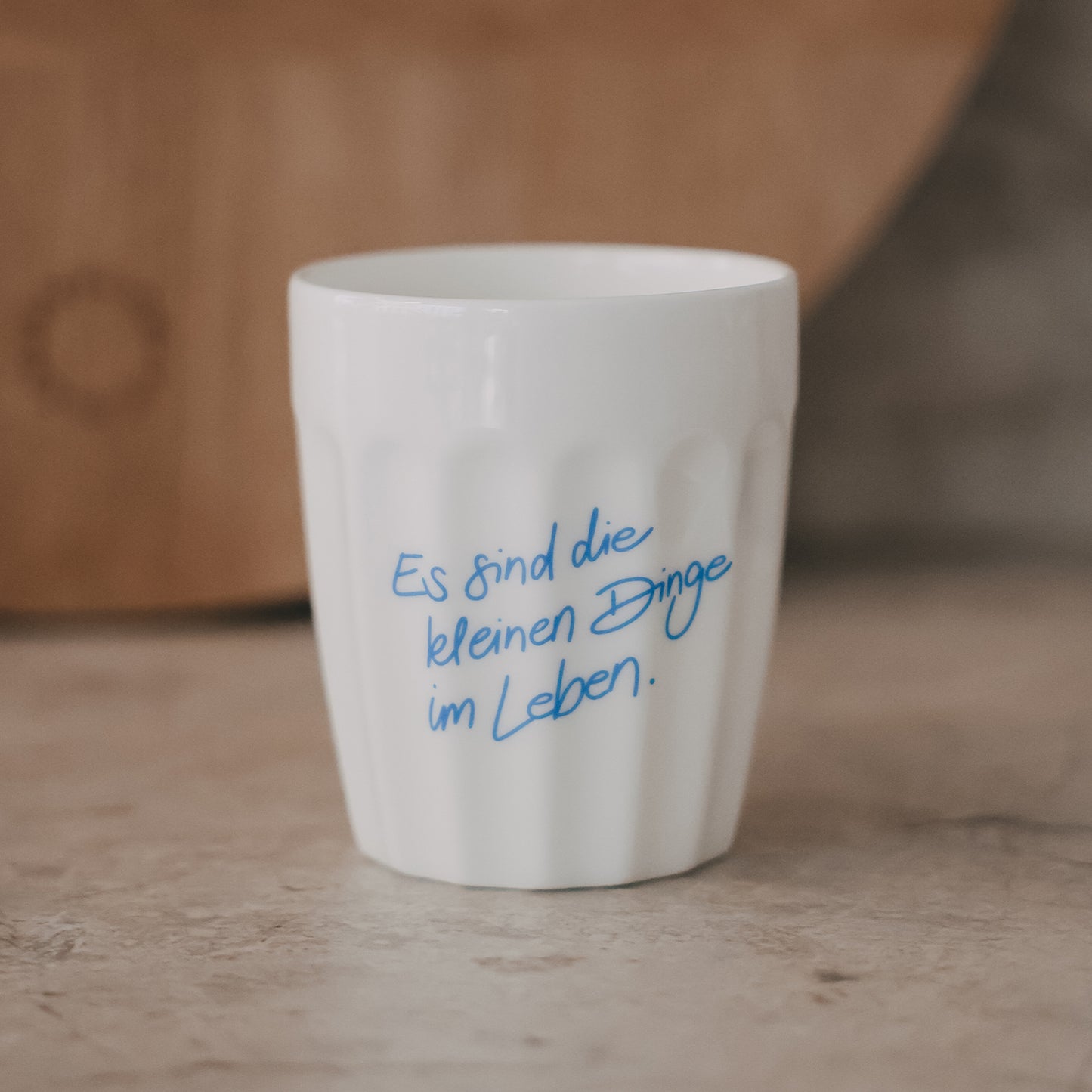 Weißer Keramikbecher mit Schriftzug „Es sind die kleinen Dinge im Leben“ in Hellblau auf natürlichem Untergrund