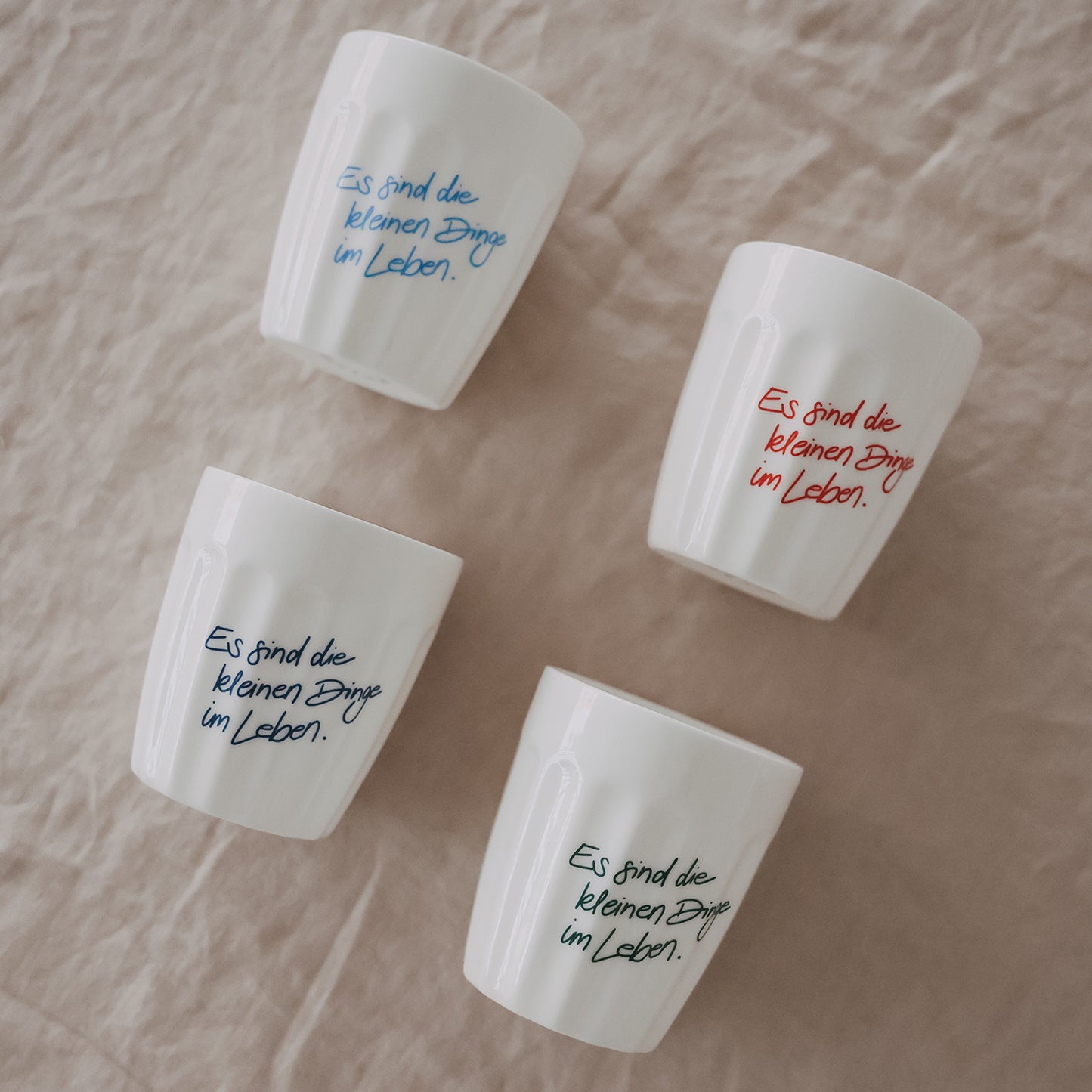 Vier weiße Becher mit Spruch „Es sind die kleinen Dinge im Leben“ locker auf Stoff drapiert, verschiedene Farben