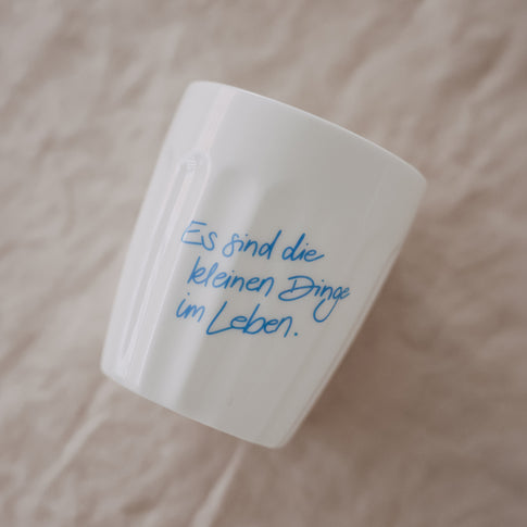 Weißer Keramikbecher mit Spruch „Es sind die kleinen Dinge im Leben“ in Hellblau, schräg liegend auf Stoff