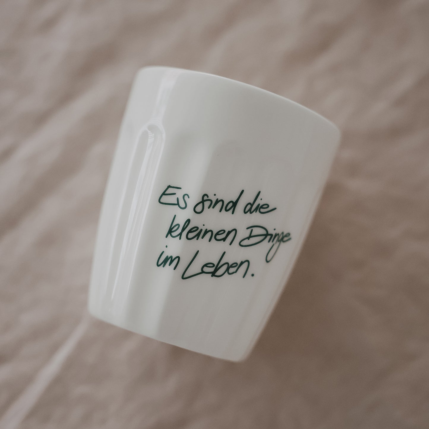 Weißer Keramikbecher mit grünem Schriftzug „Es sind die kleinen Dinge im Leben“, schräg auf Stoff platziert