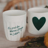 Zwei weiße Keramikbecher mit Spruch und grünem Herz, dekoriert mit Pasta und Gemüse auf hellem Stoff