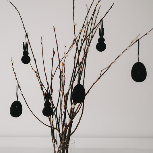 Schwarze Papier-Osteranhänger in Eiform und Hasenform an dekorierten Zweigen.
