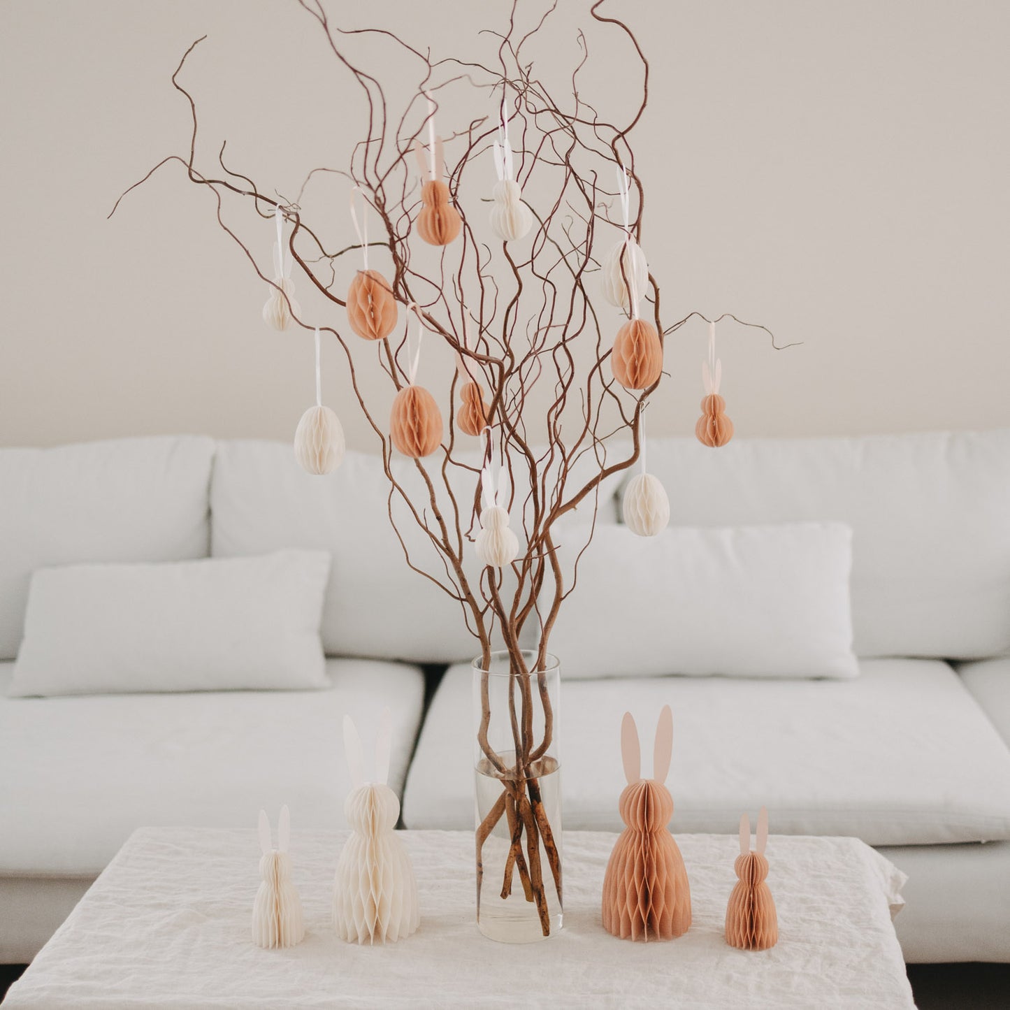 Osterstrauß mit peachfarbenen und weißen Papieranhängern in Ei- und Hasenform im Wohnzimmer.