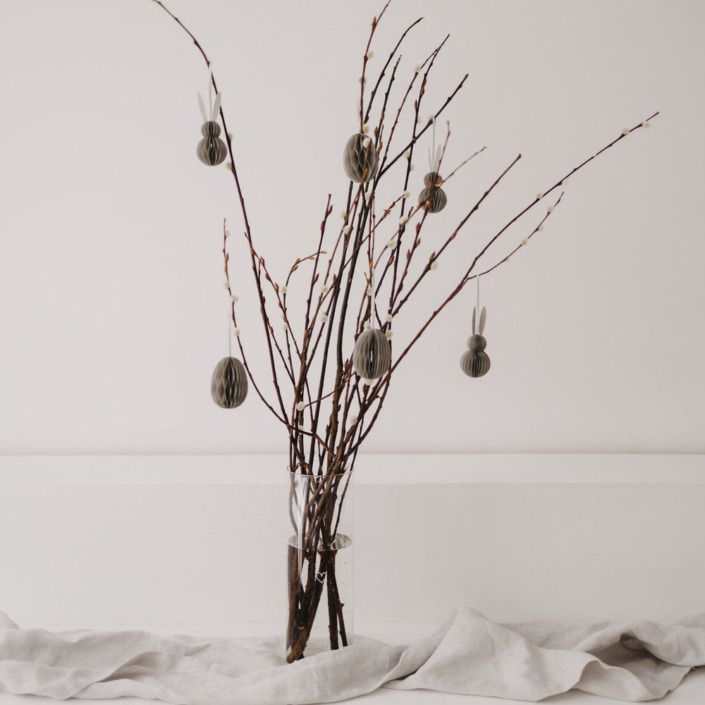 Graue Papieranhänger in Ei- und Hasenform hängen an Zweigen in einer Glasvase als Osterdeko.