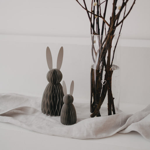 Papierhasen in Grau als Deko, zwei Größen, auf Stofftuch neben Vase mit Zweigen arrangiert.