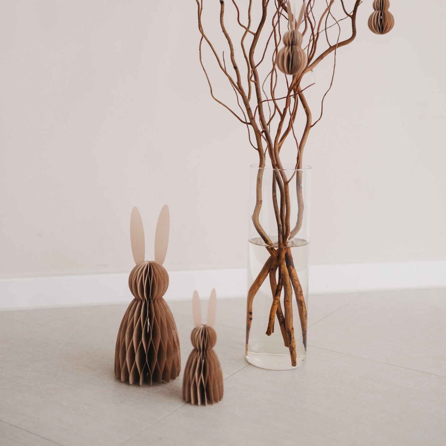 Zwei Papierhasen in Naturfarbe vor Glasvase mit Ästen als dezente Osterdeko auf dem Boden.