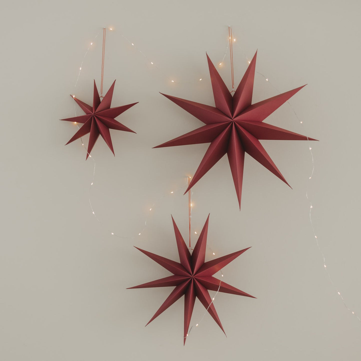 Drei rote Papiersterne in verschiedenen Größen mit warmer Lichterkette an heller Wand