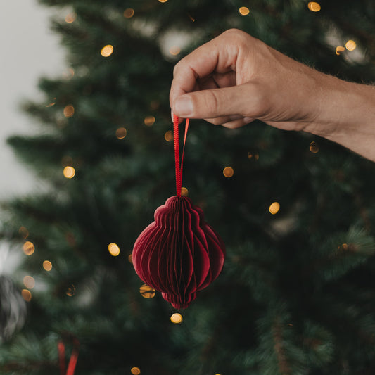 Roter Papieranhänger in Kugelform wird vor einem beleuchteten Weihnachtsbaum gehalten