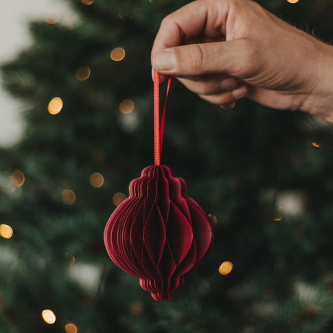 Roter Papieranhänger in Kugelform mit rotem Band vor einem festlich beleuchteten Weihnachtsbaum