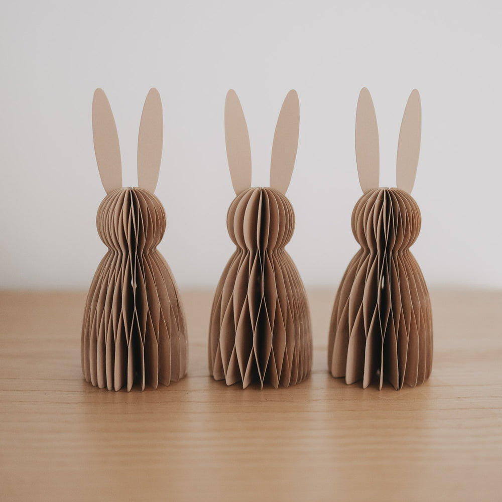 Drei naturfarbene Papierhasen in Wabenoptik als Osterdeko auf einer Holzfläche arrangiert.
