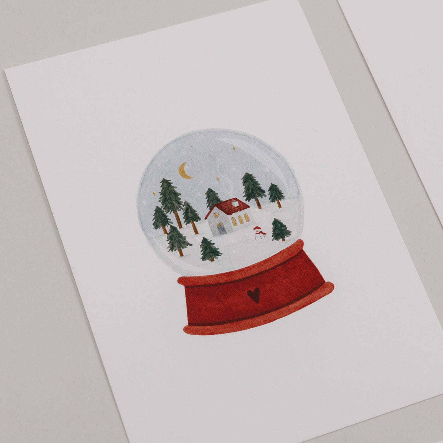 Weihnachtskarte mit Schneekugel, Häuschen, Bäumen und Mond auf rotem Sockel