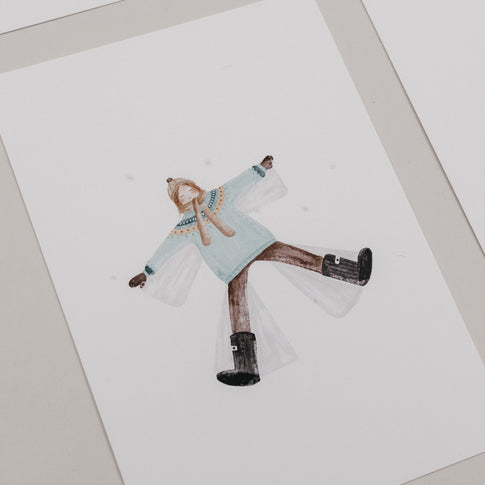 Weihnachtskarte mit Frau im Winteroutfit, die auf dem Rücken liegt und einen Schneeengel macht