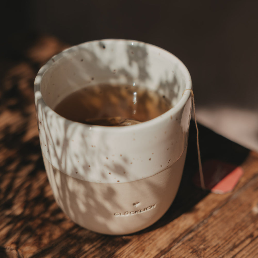 Nahaufnahme eines mit Tee gefüllten Keramikbechers „Glücklich“ auf einem rustikalen Holztisch mit warmem Licht- und Schattenspiel.