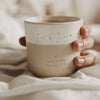 Keramikbecher „Hab einen tollen Tag“ in Beige und Weiß mit Kaffee, gemütlich im Bett gehalten