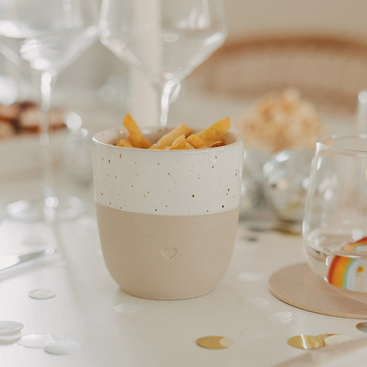 Keramikbecher mit Herzprägung und Pommes auf einem festlich gedeckten Tisch mit Konfetti und Gläsern.