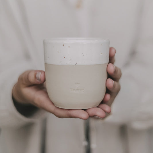 Hand hält einen schlichten, beigen Keramikbecher mit cremeweißer Glasur am oberen Rand. Auf der Vorderseite ist ein kleines Herz und darunter das Wort ‚TANTE‘ eingeprägt. Der Becher wird in beiden Händen vor dem Körper präsentiert – stilvoll, minimalistisch und mit liebevoller Botschaft, ideal als Geschenk für die Tante.