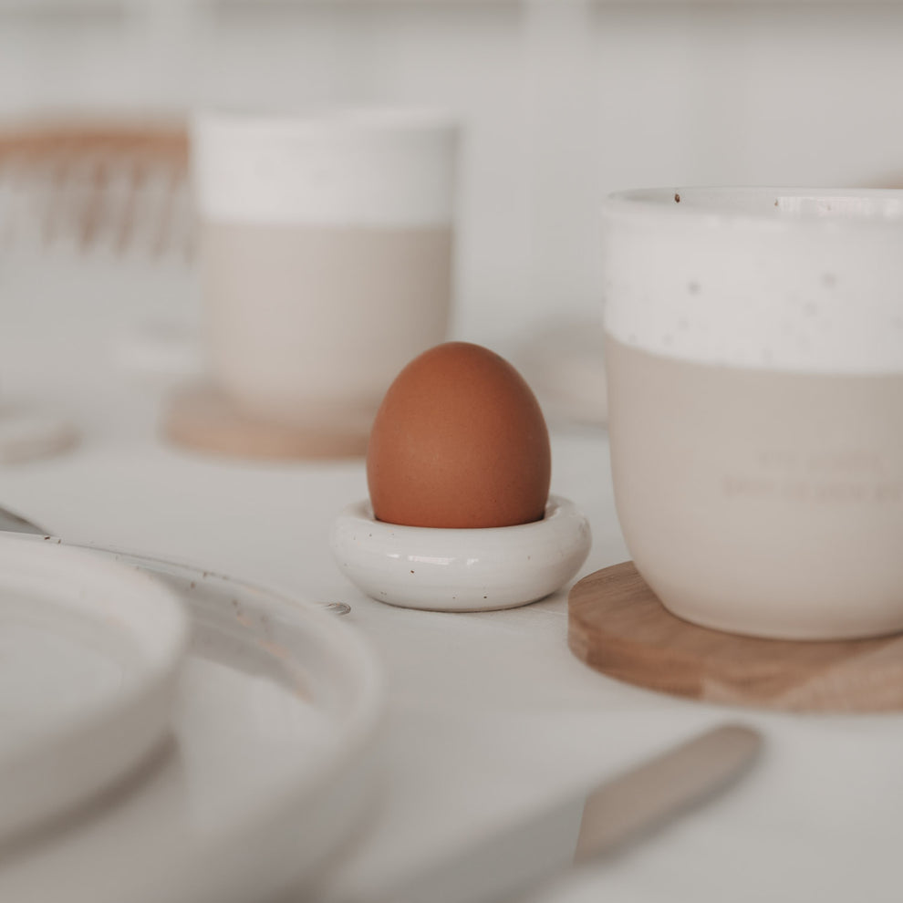 Eierbecher Calma mit Ei auf Frühstückstisch, im Hintergrund zwei Becher auf Holzuntersetzern