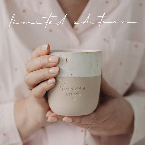 Person hält beige-weißen Limited-Edition-Becher mit Herzensmensch-Gravur nah vor dem Körper in gemütlicher Atmosphäre.