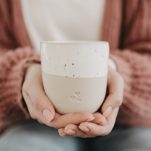 Person hält den beigen Keramikbecher mit Herzprägung und gesprenkelter Glasur sanft in beiden Händen.