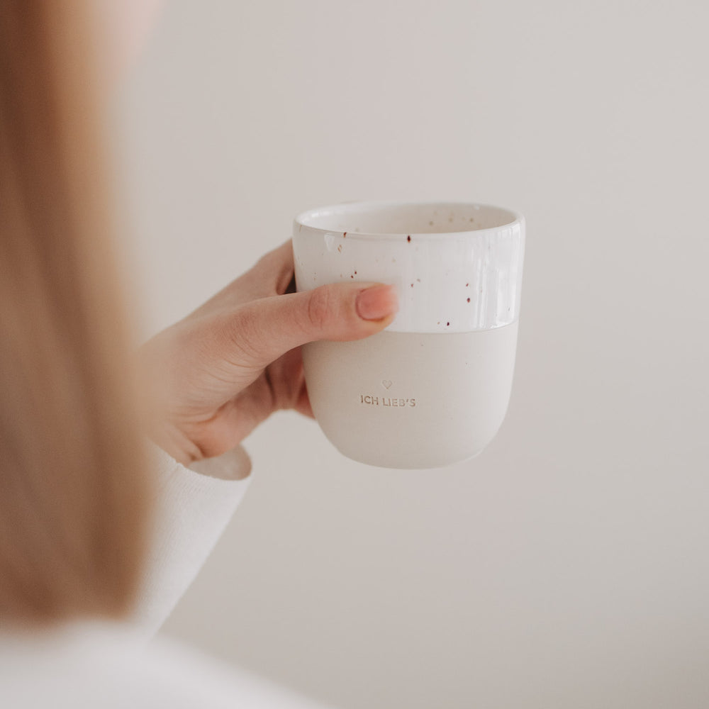 Seitliche Aufnahme einer Hand mit der beige-weißen „Ich lieb’s“-Tasse mit Sprenkeln.