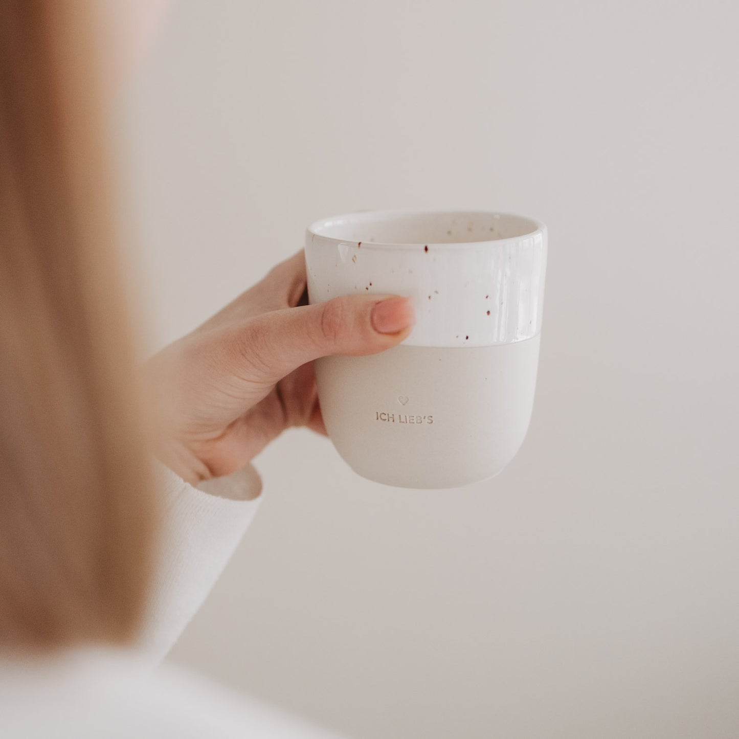 Seitliche Aufnahme einer Hand mit der beige-weißen „Ich lieb’s“-Tasse mit Sprenkeln.
