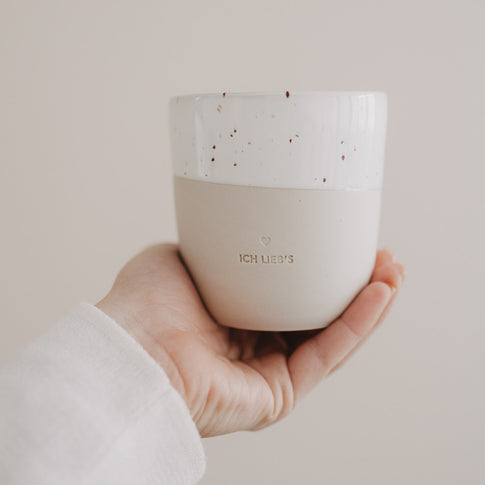 Hand hält beige-weiße „Ich lieb’s“-Tasse mit Sprenkelglasur vor hellem Hintergrund.