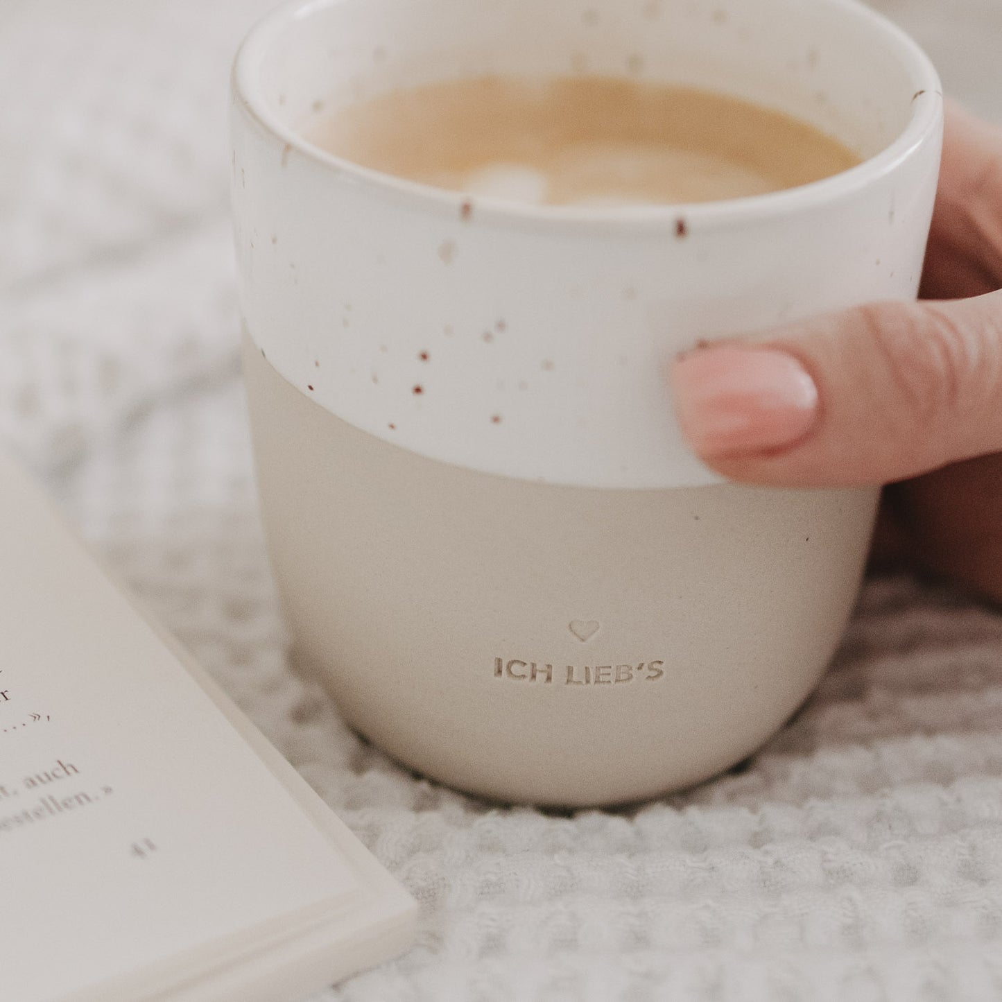 „Ich lieb’s“-Tasse mit Sprenkelglasur in der Hand, gefüllt mit Kaffee, neben Buch.