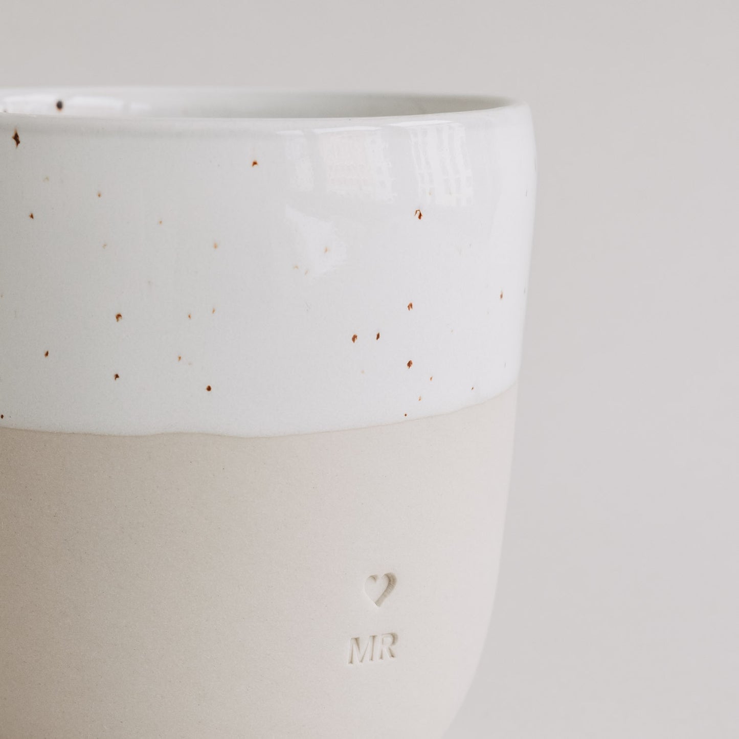 Seitliche Nahaufnahme des MR-Bechers mit Herz- und MR-Prägung auf matter beiger Keramik.