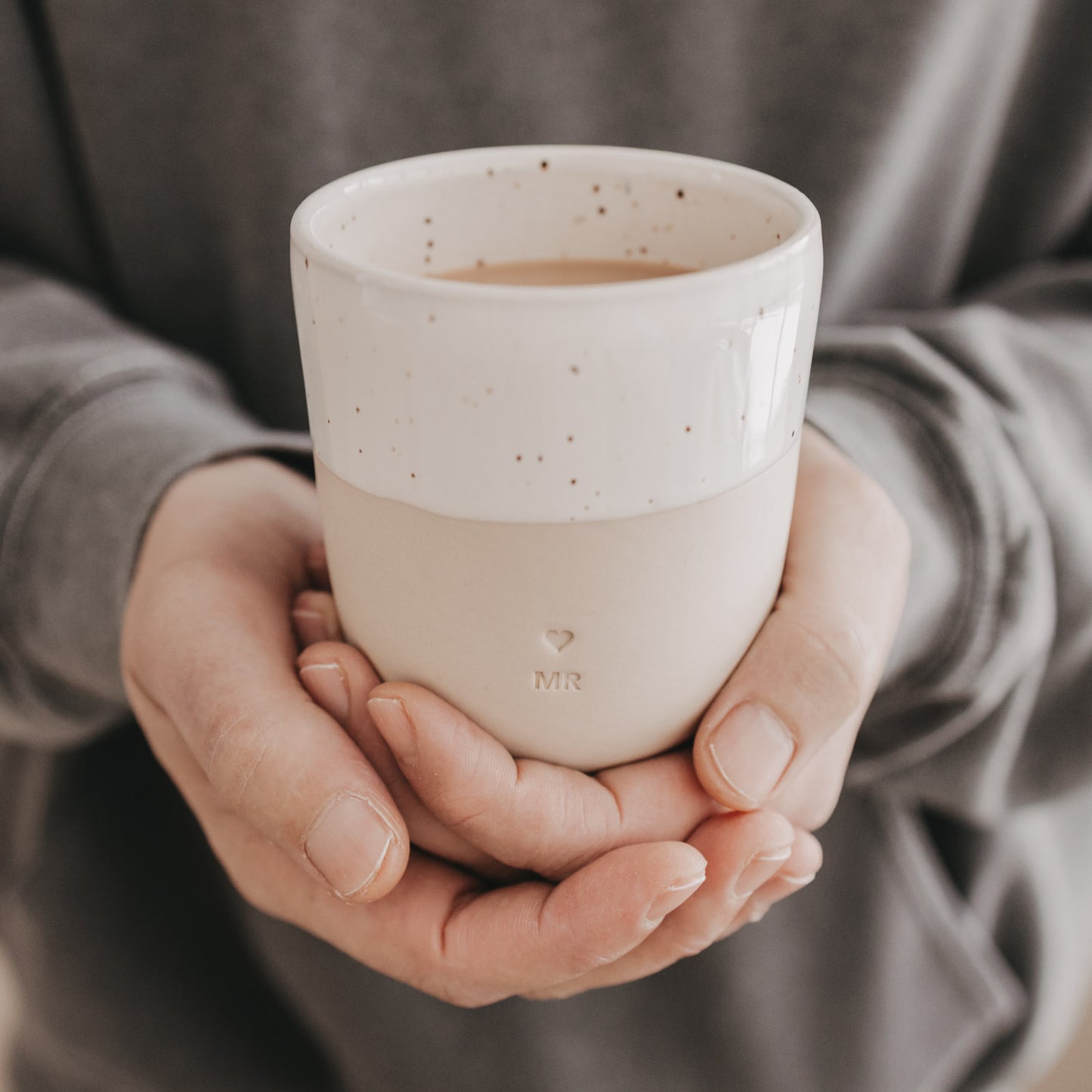 Person hält den MR-Becher mit weißer Sprenkelglasur und beigefarbener Keramik in beiden Händen.