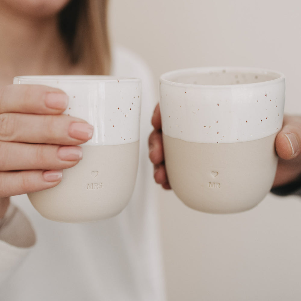 Zwei Personen halten passende Keramikbecher mit MR- und MRS-Prägung in weißer Sprenkelglasur.