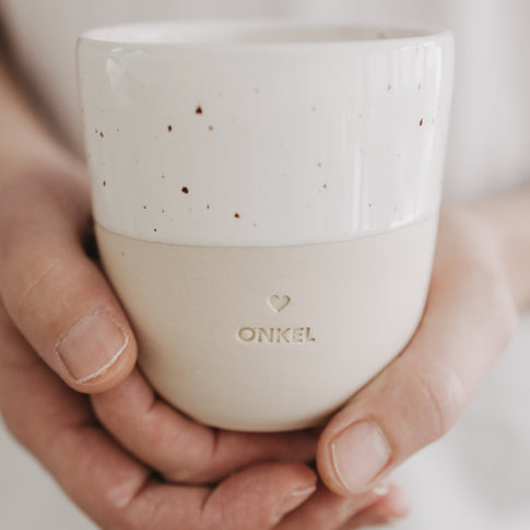 Keramikbecher ONKEL in Beige-Weiß mit Speckle-Glasur, wird in beiden Händen gehalten.