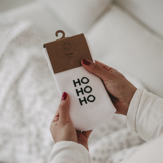 Nahaufnahme einer Person, die ein Paar weiße Socken mit schwarzem Schriftzug ‚HO HO HO‘ in der Verpackung hält. Im Hintergrund ist eine helle Bettdecke zu sehen.