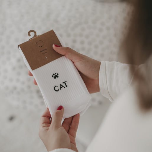 Eine Person in weißem Oberteil hält ein Paar weiße Socken mit einem schwarzen Pfotenabdruck und dem Wort „CAT“ in der Hand; die Socken sind mit einer braunen Kartonaufhängung verpackt.