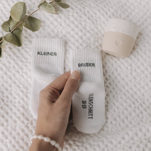 Hand hält Babysocken „Kleiner Bruder“ auf weißer Decke mit Eukalyptuszweig