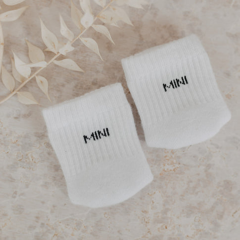 Zwei „MINI“-Babysocken auf beigem Untergrund neben dekorativen Trockenblättern arrangiert