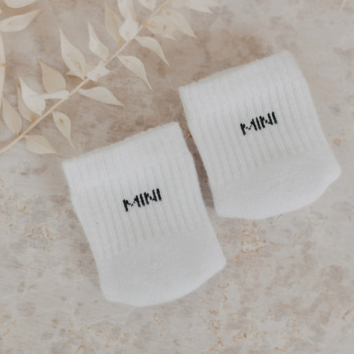Zwei „MINI“-Babysocken auf beigem Untergrund neben dekorativen Trockenblättern arrangiert