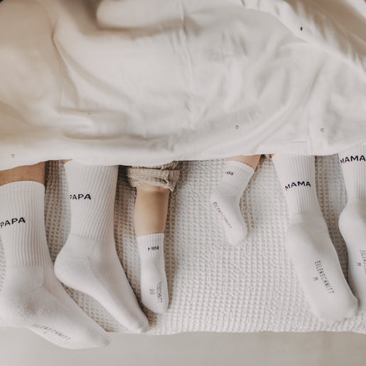 Familie mit „MAMA“, „PAPA“ und „MINI“-Socken liegt gemütlich unter einer weißen Decke im Bett