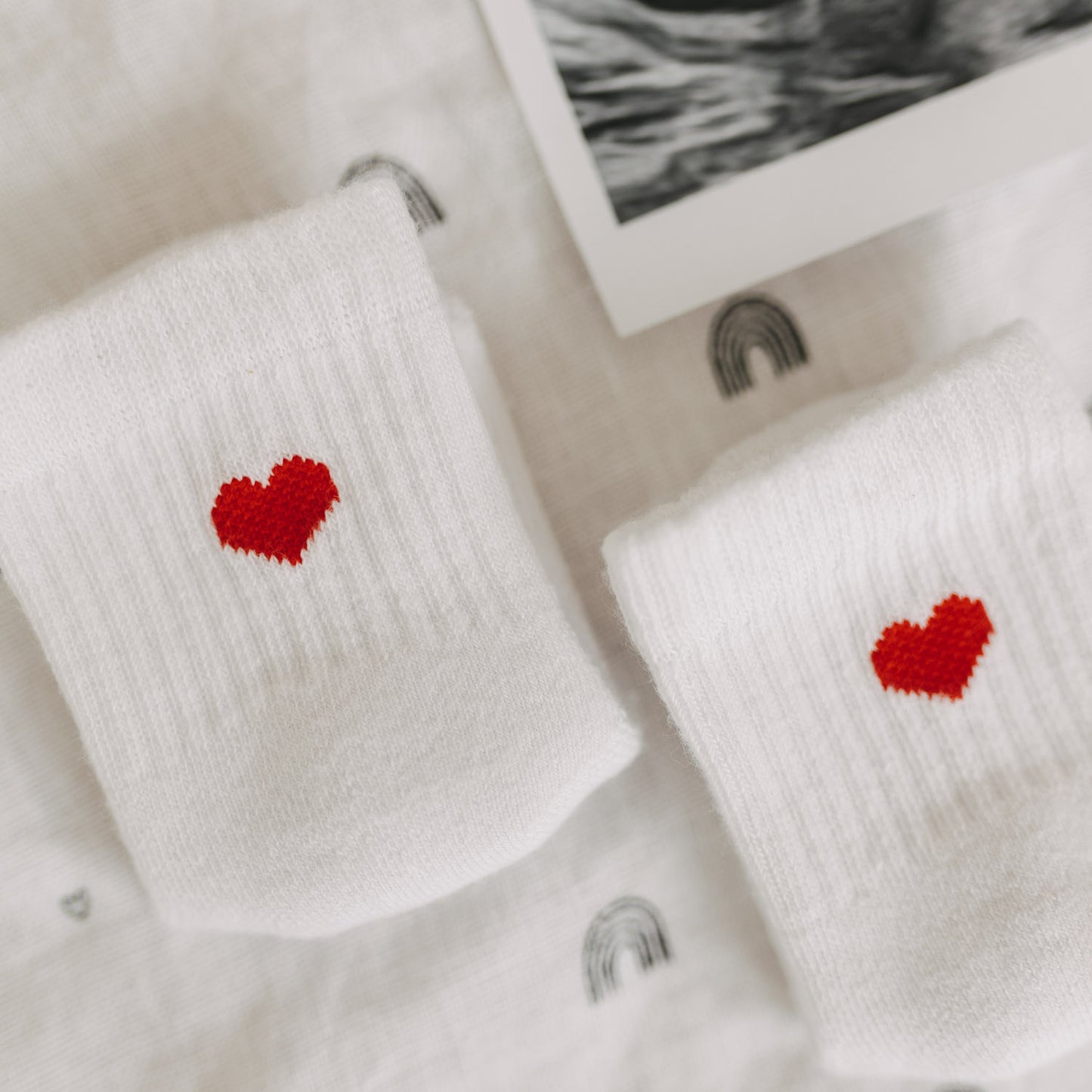 Nahaufnahme der roten Herzmotive auf weißen Babysocken auf Stoff mit Regenbogenmuster