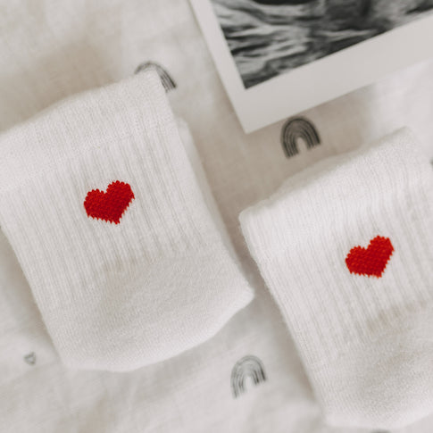 Nahaufnahme der roten Herzmotive auf weißen Babysocken auf Stoff mit Regenbogenmuster