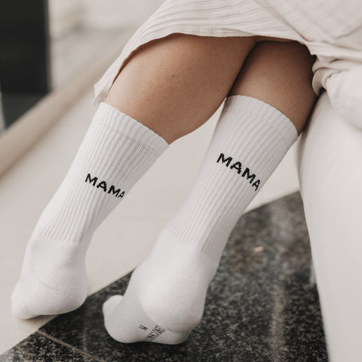 Frau sitzt entspannt mit übergeschlagenen Beinen und trägt weiße Socken mit Mama-Schriftzug.