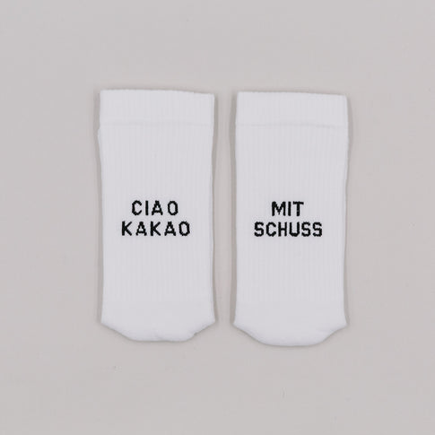 Weiße Socken mit schwarzer Stickerei „Ciao Kakao“ und „Mit Schuss“ auf neutralem Untergrund nebeneinander liegend