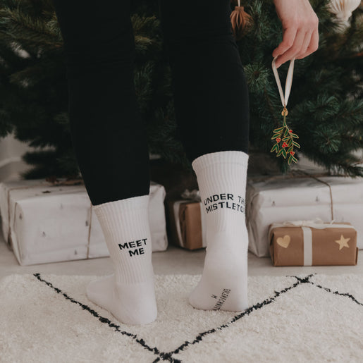 Rückansicht einer Person in den Mistletoe-Socken mit Mistelzweig in der Hand vor weihnachtlich geschmücktem Baum