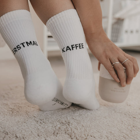Weiße Socken „Erstmal Kaffee“ getragen im Alltag mit Tasse in der Hand