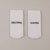 Flachliegende weiße Socken mit „Erstmal Kaffee“ Schriftzug minimalistisch