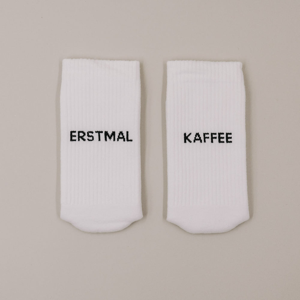 Flachliegende weiße Socken mit „Erstmal Kaffee“ Schriftzug minimalistisch