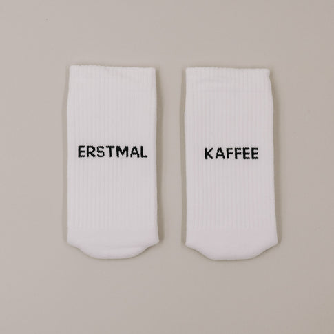Flachliegende weiße Socken mit „Erstmal Kaffee“ Schriftzug minimalistisch