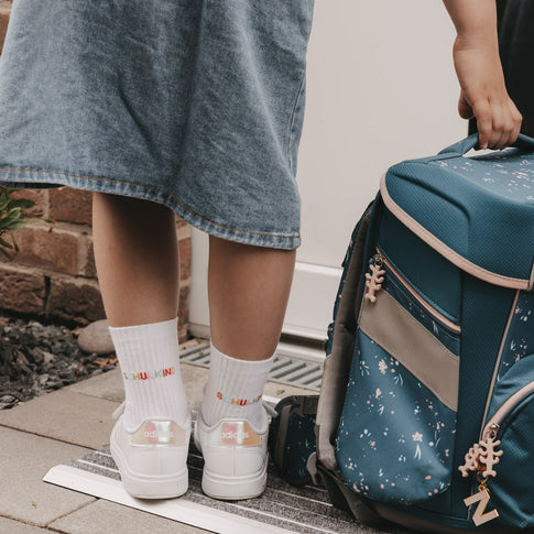Kind mit Schulranzen trägt weiße Socken „Schulkind“ und Sneaker vor der Haustür