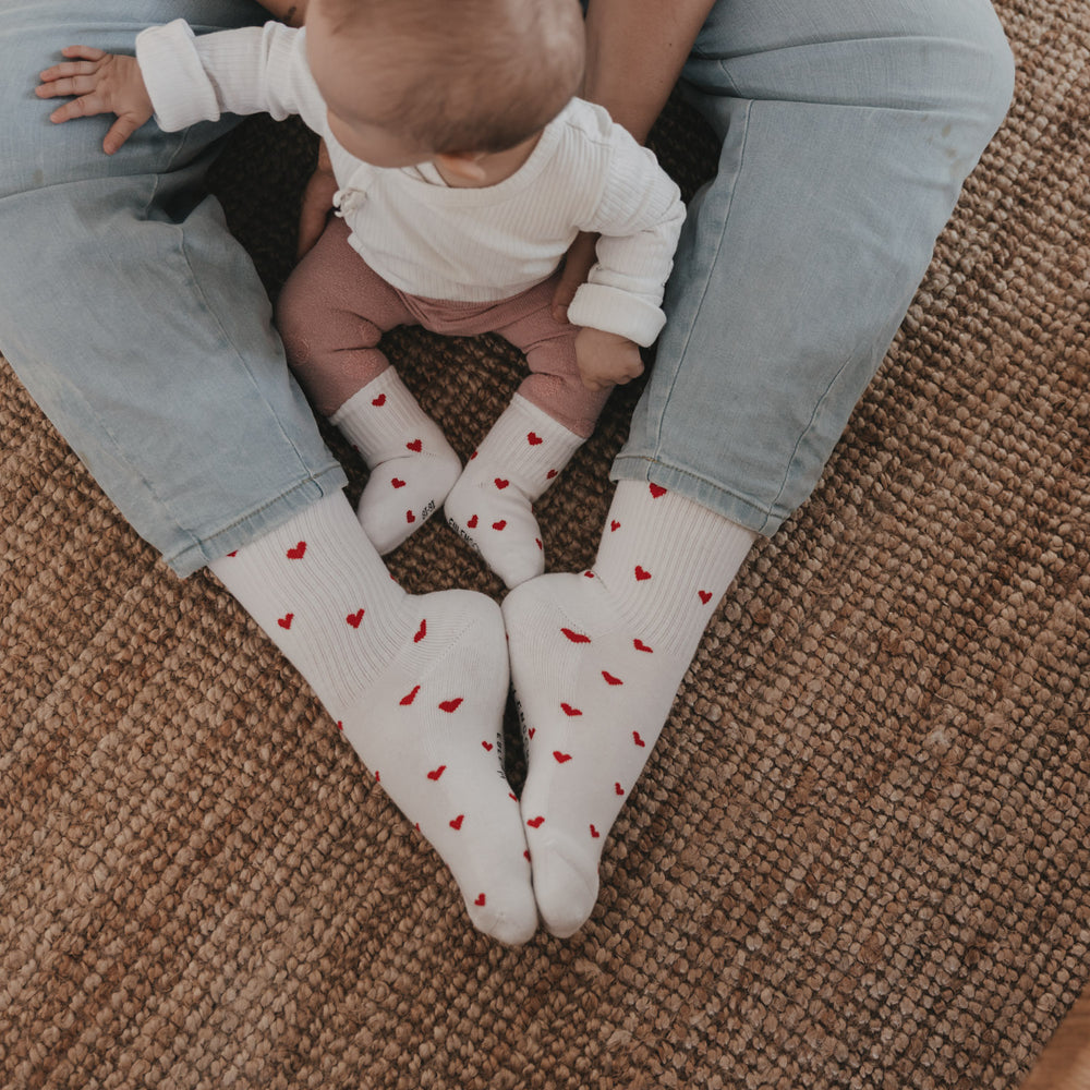 Familie trägt passende Socken mit roten Herzen, Baby sitzt zwischen Erwachsenen auf Teppich