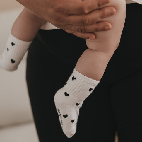 Baby wird gehalten, trägt weiße Herzsocken mit schwarzen Punkten, weiches Material