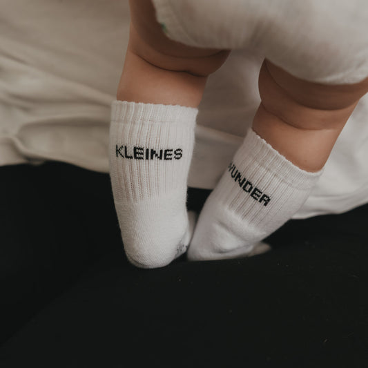 Baby trägt weiße Socken mit Schriftzug „Kleines Wunder“, Füße auf dunklem Untergrund